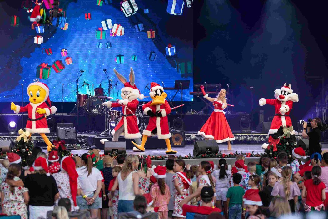 Families enjoying Christmas in Gold Coast at a lively stage show featuring festive characters in Santa costumes performing for children under colourful lights.